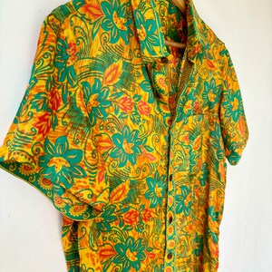 May include: A short-sleeved button-down shirt with a floral print in shades of yellow, green, and orange. The shirt has a pocket on the left side and a label that reads "Jaipur Craft and Art".
