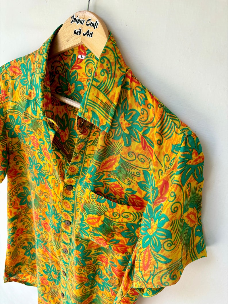 May include: A yellow, green and orange short-sleeved shirt with a floral pattern. The shirt has a button-up front and a pocket on the left side. The shirt is hanging on a wooden hanger with the text "Jaipur Craft and Art" printed on it.