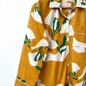 Puede incluir: Una camisa de manga larga con fondo amarillo mostaza y estampado floral abstracto blanco y verde. La camisa tiene un cierre de botones en la parte delantera y un cuello.