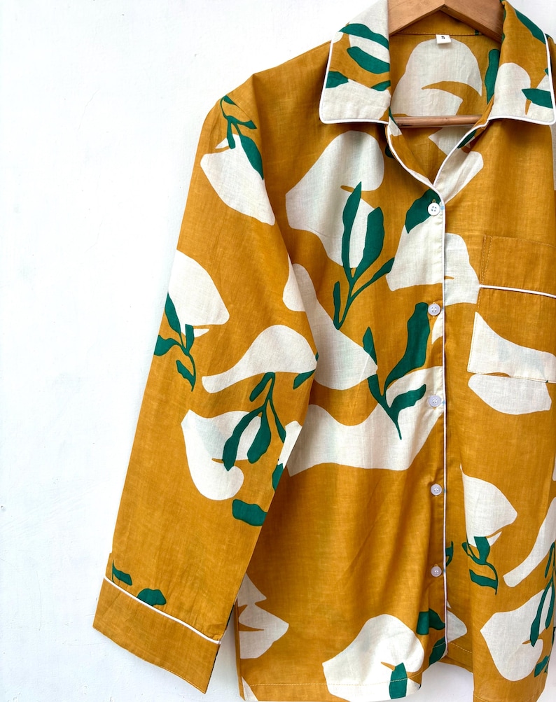 Puede incluir: Una camisa de manga larga con fondo amarillo mostaza y estampado floral abstracto blanco y verde. La camisa tiene un cierre de botones en la parte delantera y un cuello.