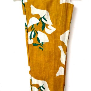 Puede incluir: Pantal&oacute;n amarillo mostaza con estampado floral blanco y verde. El pantal&oacute;n est&aacute; colgado en una percha de madera.