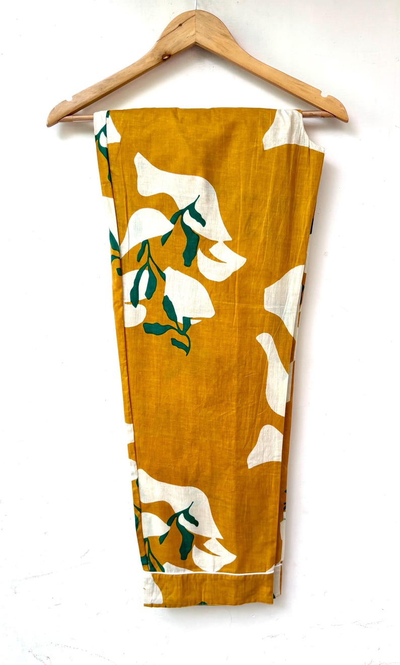 Puede incluir: Pantal&oacute;n amarillo mostaza con estampado floral blanco y verde. El pantal&oacute;n est&aacute; colgado en una percha de madera.