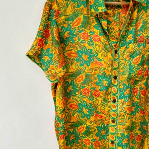 May include: A yellow, green and orange short-sleeved button-down shirt with a floral print. The shirt has a pocket on the left side and a button closure.