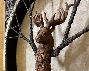 Può includere: Una scultura in legno di una testa di cervo con corna, montata su una cornice circolare con cinghie di cuoio intrecciate. Il cervo ha un design dettagliato e realistico ed è dipinto in tonalità marroni.