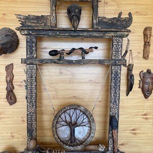 May include: A wooden frame with the words "Open your drum" carved into the top. A round drum with a tree of life design hangs from the frame. The frame is decorated with carvings and has a variety of other items hanging from it, including a skull, a snake, and a feather.
