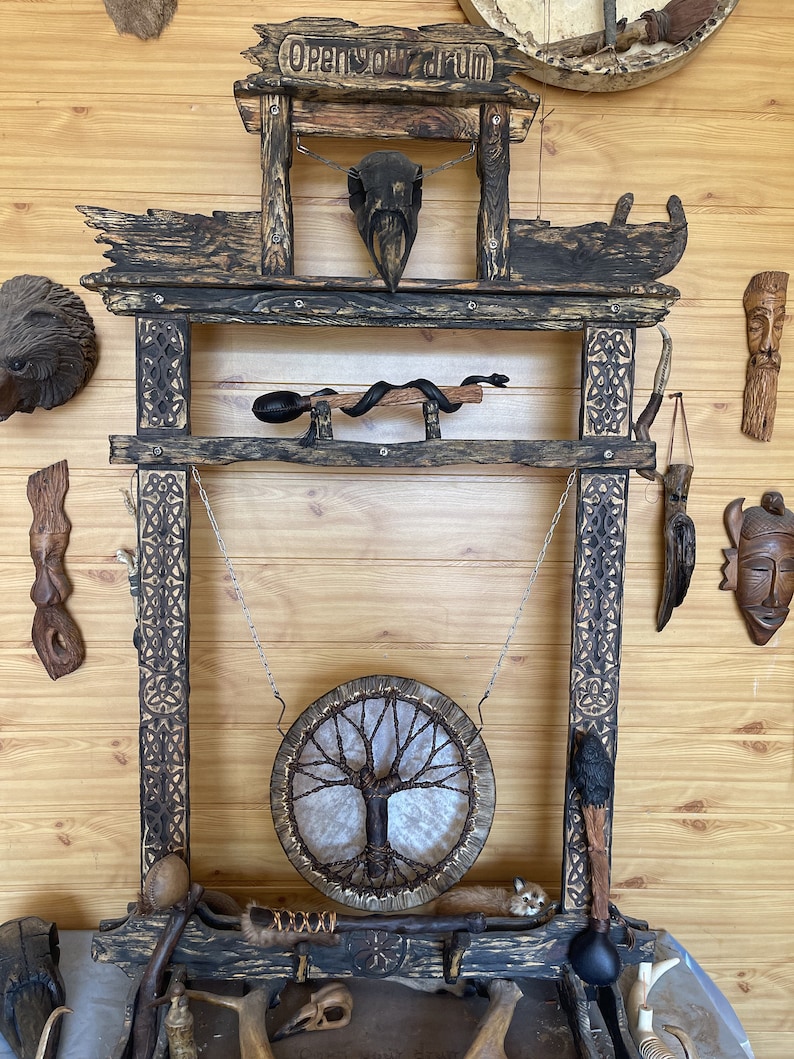 May include: A wooden frame with the words "Open your drum" carved into the top. A round drum with a tree of life design hangs from the frame. The frame is decorated with carvings and has a variety of other items hanging from it, including a skull, a snake, and a feather.