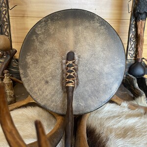 May include: A brown leather drum with a wooden handle and a fringe of brown fur. The drum is resting on a white fur surface.