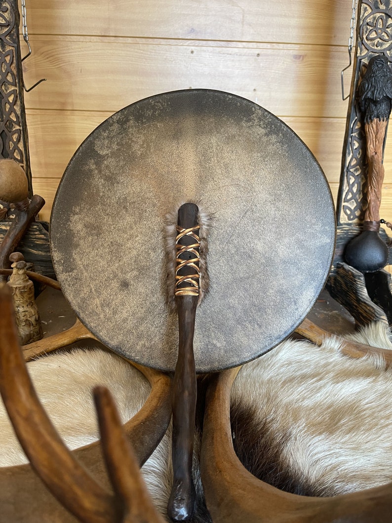 May include: A brown leather drum with a wooden handle and a fringe of brown fur. The drum is resting on a white fur surface.