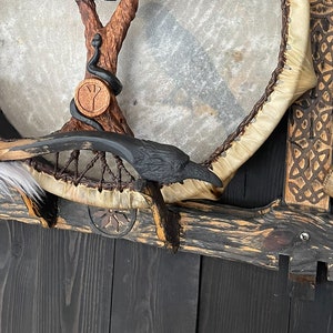 May include: A wooden frame with a circular drum, a carved wooden tree with a snake wrapped around it, a black raven, and a small leather-covered ball on a stick. The frame is decorated with intricate carvings.