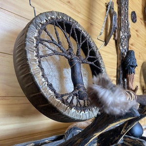 May include: A brown leather drum with a tree of life design hanging from a chain. The drum is made of wood and has a natural leather head. The tree of life design is made of woven brown cord.