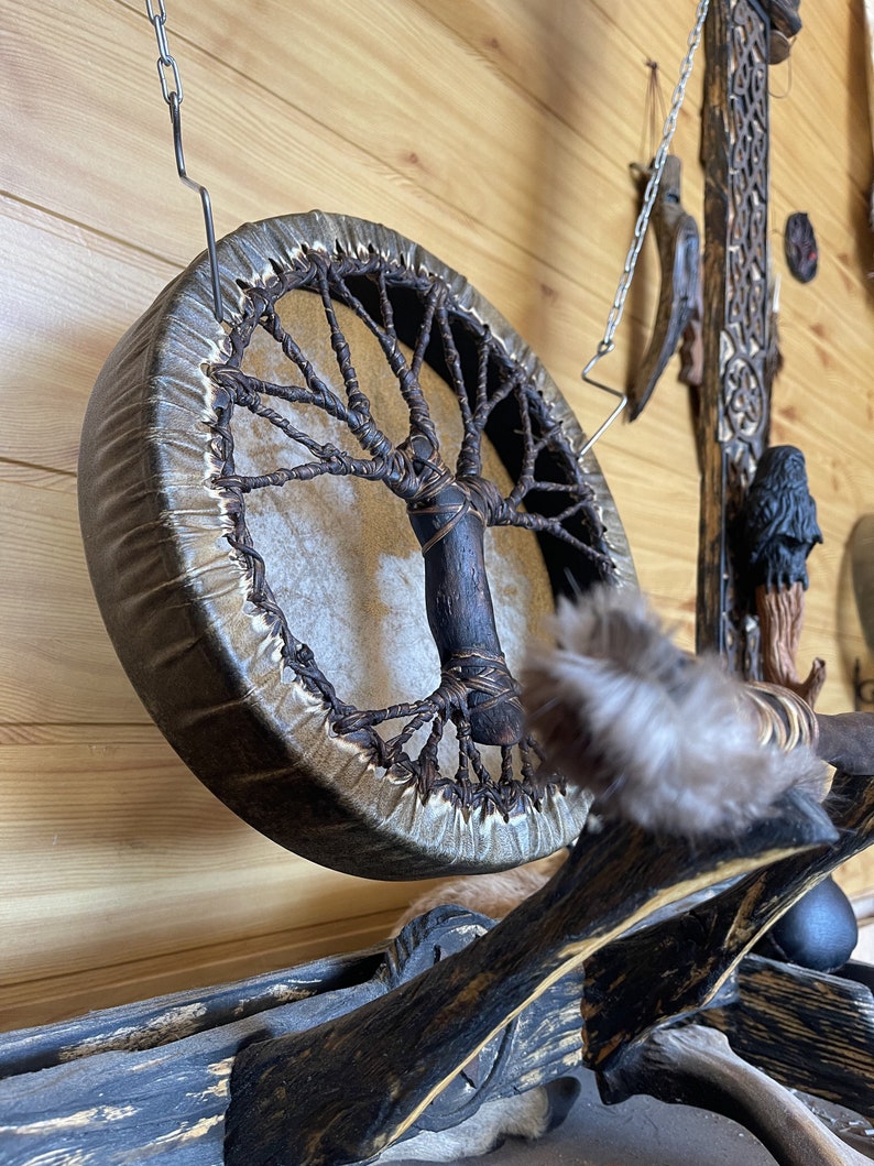 May include: A brown leather drum with a tree of life design hanging from a chain. The drum is made of wood and has a natural leather head. The tree of life design is made of woven brown cord.