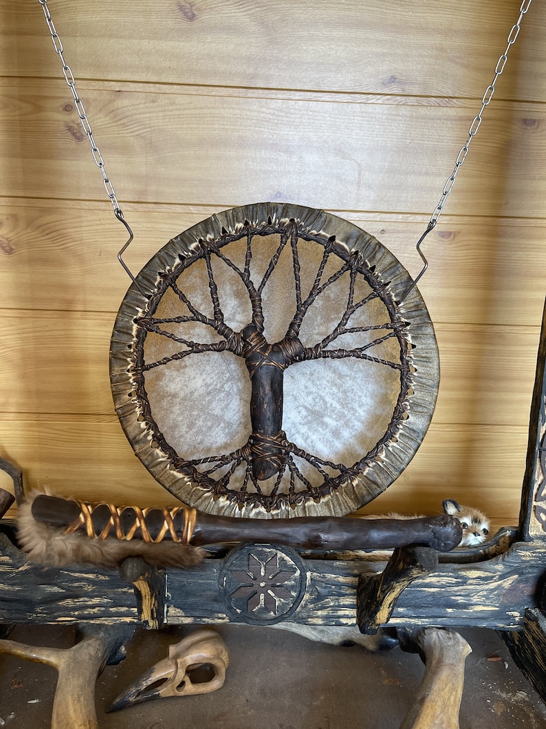 May include: A brown leather drum with a tree of life design hanging from a chain. The drum is made of wood and has a brown leather strap. The drum is hanging on a wooden wall.