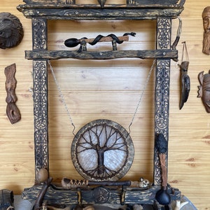 May include: A wooden frame with a carved design, featuring a hanging drum with a tree of life design, a snake, a skull, and other wooden objects. The frame is decorated with a black and brown finish.