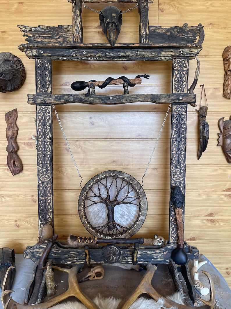 May include: A wooden frame with a carved design, featuring a hanging drum with a tree of life design, a snake, a skull, and other wooden objects. The frame is decorated with a black and brown finish.