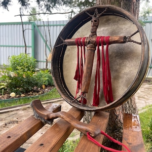 Pode incluir: Um tambor de moldura de madeira com uma cabeça de couro branco e uma figura de madeira com um rosto vermelho e tecido vermelho pendurado em seus braços. Um batedor de madeira com uma escova em uma extremidade está sentado em um banco de madeira.
