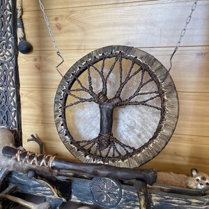 May include: A brown leather drum with a tree of life design hanging from a chain on a wooden wall. The drum is surrounded by other wooden objects, including a small animal figurine.