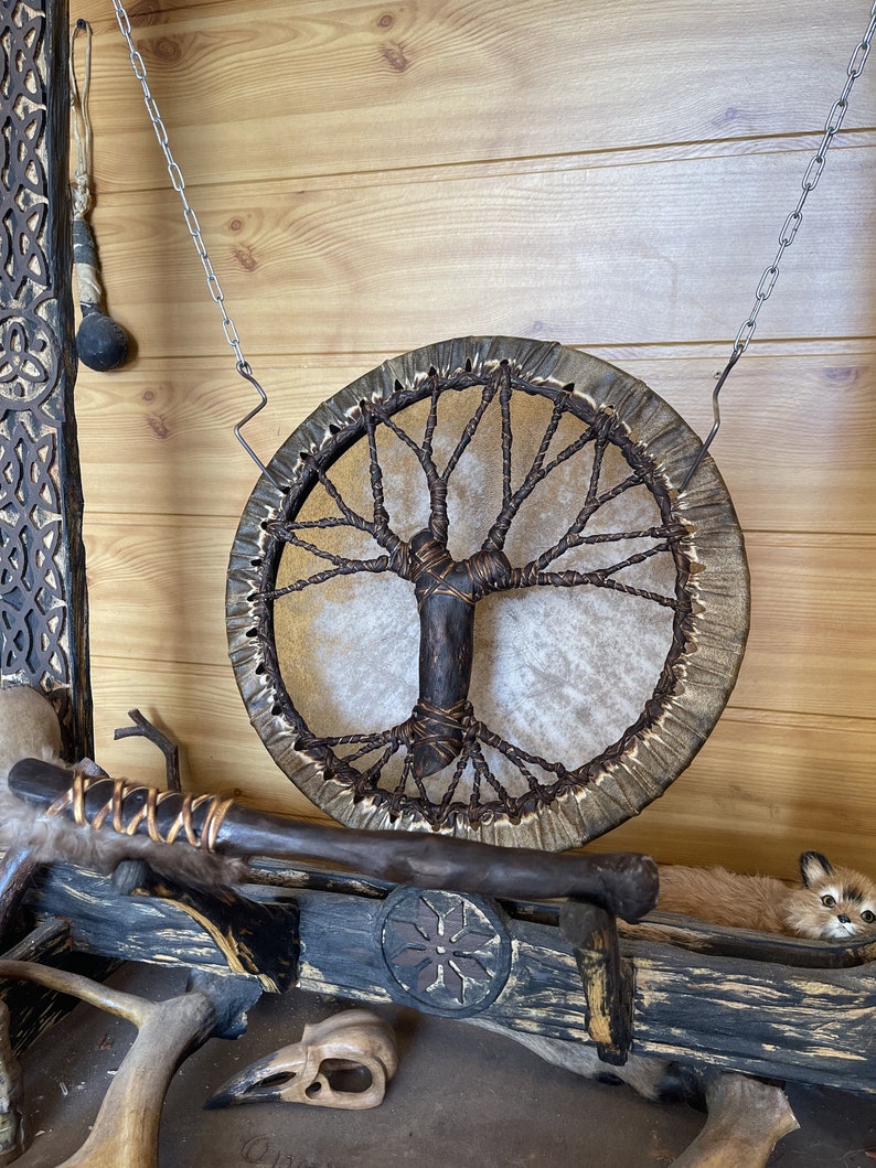 May include: A brown leather drum with a tree of life design hanging from a chain on a wooden wall. The drum is surrounded by other wooden objects, including a small animal figurine.