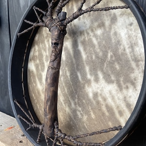 May include: A handcrafted drum with a black frame and a natural leather drumhead. The drum features a unique tree-shaped handle made of wood with a carved face.