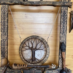 May include: A wooden frame with intricate carvings, holding a hanging drum with a tree of life design. The drum is made of leather and has a natural wood frame. The frame also holds various other items, including a small animal figurine, a wooden staff, and a leather-wrapped stick.