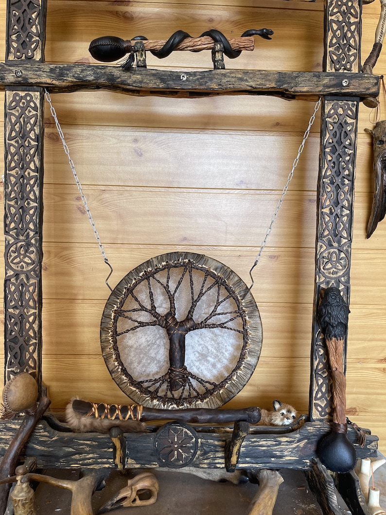 May include: A wooden frame with intricate carvings, holding a hanging drum with a tree of life design. The drum is made of leather and has a natural wood frame. The frame also holds various other items, including a small animal figurine, a wooden staff, and a leather-wrapped stick.