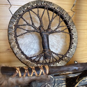 May include: A brown leather drum with a tree of life design woven into the leather. The drum is hanging from a chain and has a wooden handle.