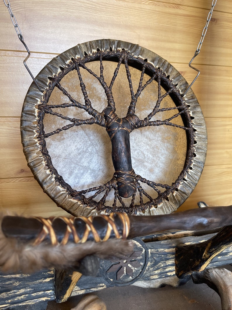 May include: A brown leather drum with a tree of life design woven into the leather. The drum is hanging from a chain and has a wooden handle.