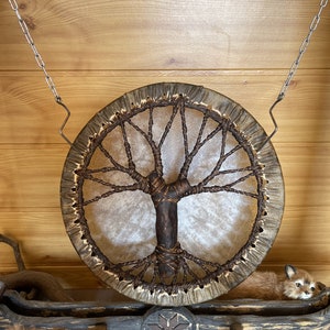 May include: A brown leather drum with a tree of life design hanging from a chain on a wooden wall. A small stuffed fox is lying on a wooden shelf below the drum.