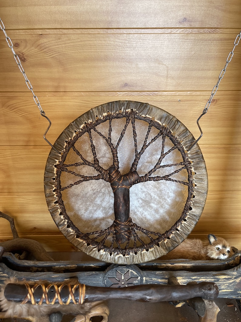 May include: A brown leather drum with a tree of life design hanging from a chain on a wooden wall. A small stuffed fox is lying on a wooden shelf below the drum.