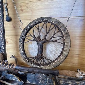 May include: A brown leather drum with a tree of life design hanging from a chain on a wooden wall. The drum is made of natural materials and has a rustic look.