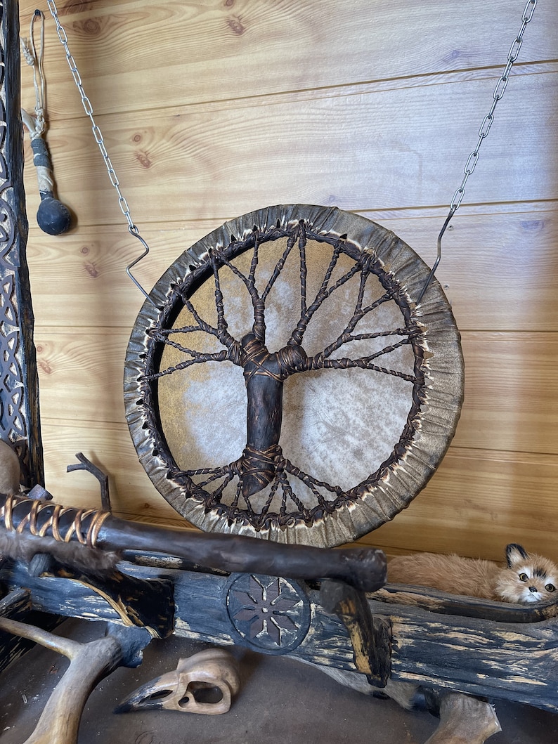 May include: A brown leather drum with a tree of life design hanging from a chain on a wooden wall. The drum is made of natural materials and has a rustic look.