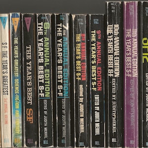 Op de afbeelding: Een verzameling van 12 paperback boeken, allemaal met zwarte omslagen en witte tekst. De boeken zijn getiteld "The Year's Best S-F" en zijn genummerd van de 6e tot de 12e jaarlijkse editie. De boeken zijn allemaal bewerkt door Judith Merril.