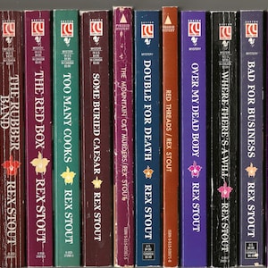 May include: A collection of 17 paperback mystery novels by Rex Stout featuring the detective Nero Wolfe. The books are arranged vertically in a row and feature colorful covers with titles such as "The Red Box", "The Rubber Band", "Some Buried Caesar", and "Double for Death".