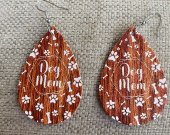 Puede incluir: Un par de pendientes de madera marrón con un diseño de huella de pata y hueso blanco. Los pendientes tienen ganchos plateados y dicen "Dog Mom" en texto blanco.