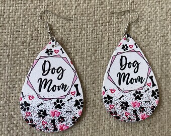 Puede incluir: Un par de pendientes blancos en forma de lágrima con un diseño rosa y negro. Los pendientes presentan el texto "Dog Mom" con huellas de patas y corazones.