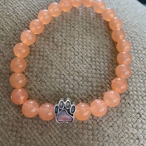 May include: A peach-colored beaded bracelet with a silver paw print charm.