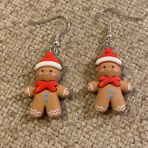 May include: A pair of gingerbread man earrings with red hats, white trim, red bows, and blue buttons. The earrings are made of polymer clay and have silver hooks.