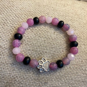 May include: A beaded bracelet featuring a mix of pink, black, and light pink beads. The bracelet has a silver-colored paw print charm in the center. The beads are round and have a textured appearance.