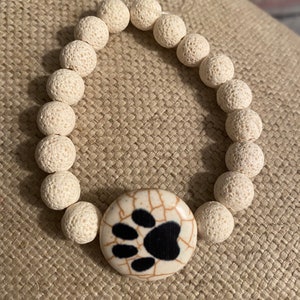 Op de afbeelding: Wit lavastenen armband met een zwarte pootprint bedel. De armband is gemaakt van natuurlijke lavastenen en heeft een eenvoudig ontwerp.
