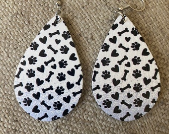 Puede incluir: Pendientes en forma de lágrima con fondo blanco y un estampado de huellas de patas negras, corazones y huesos. Los pendientes tienen ganchos para las orejas de color plateado. El diseño se repite en ambos pendientes.