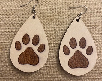 Puede incluir: Un par de pendientes de madera con un diseño de huella de pata marrón. Los pendientes tienen forma de lágrima y tienen ganchos plateados.