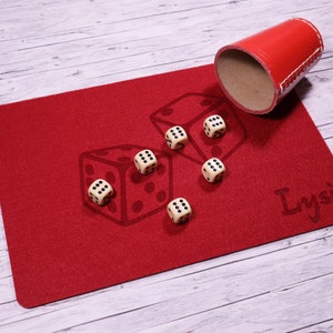 Könnte beinhalten: Ein rotes Filz-Würfeltablett mit dem Namen "Lysi" darauf gedruckt. Sechs weiße Würfel mit schwarzen Punkten sind auf dem Tablett verstreut. Ein roter Leder-Würfelbecher liegt neben dem Tablett umgedreht.