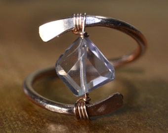 May include: A handcrafted copper ring featuring a clear, faceted gemstone. The ring has a unique, open design with the gemstone held in place by wire wrapping. The copper band curves around the finger, creating an elegant and modern look.