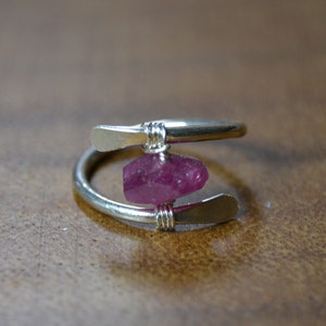 May include: A silver-colored ring with a raw, pink gemstone at its center. The ring has a spiral design, with the gemstone held in place by wire. The ring is displayed on a wooden surface.