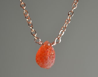 May include: A delicate necklace featuring a teardrop-shaped, orange-colored gemstone pendant. The pendant is suspended from a delicate, rose-gold colored chain. The necklace is displayed against a neutral gray background.