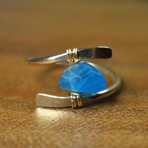 May include: A silver-toned ring with a blue, faceted gemstone pendant. The ring has a unique, open design with gold wire accents. The gemstone is a vibrant shade of blue, adding a pop of color to the jewelry.