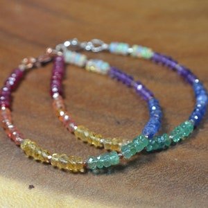 May include: Two beaded bracelets with a rainbow color scheme. The bracelets feature small, faceted gemstones in shades of red, orange, yellow, green, blue, and purple. The bracelets are displayed on a wooden surface.