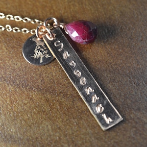 Puede incluir: Un collar dorado con una cadena, un colgante con el nombre "Savannah" grabado, un pequeño dije redondo con un diseño de piña y una piedra preciosa de color rojo oscuro. El collar se muestra sobre una superficie marrón texturizada.