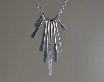 May include: A silver-toned necklace with a pendant featuring multiple vertical, textured metal bars. The bars are arranged in a fan-like pattern, suspended from a delicate chain. The necklace has a minimalist, modern design.