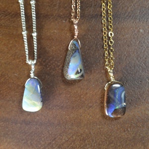 May include: Three necklaces with iridescent opal pendants. The pendants are different shapes and sizes, with silver, rose gold, and gold chains. The opals display a range of colors, including blue, green, and yellow. The background is a brown, textured surface.