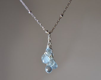 Puede incluir: Un delicado collar de plata con un colgante de cuentas de piedras preciosas azul claro. Las cuentas están facetadas, captando la luz, y cuelgan de una fina cadena de plata. El collar es una joya sencilla y elegante.